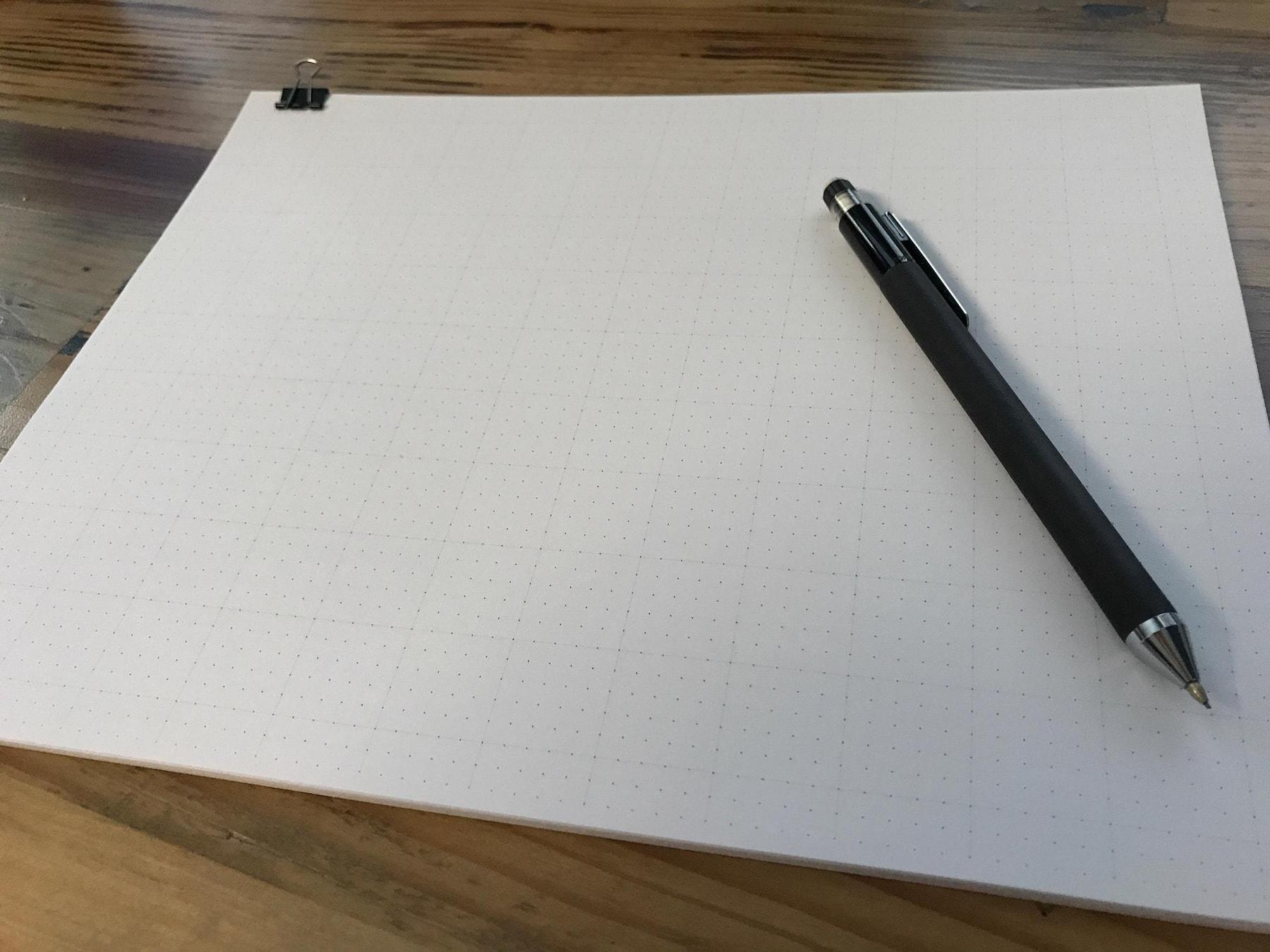 A small stack of dot paper on top of a wooden desk with a mechanical pencil on top